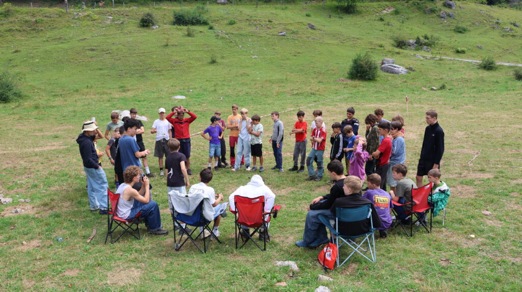Kinder und Leiter bilden im Sommerlager gemeinsam einen Kreis auf einer Wiese.