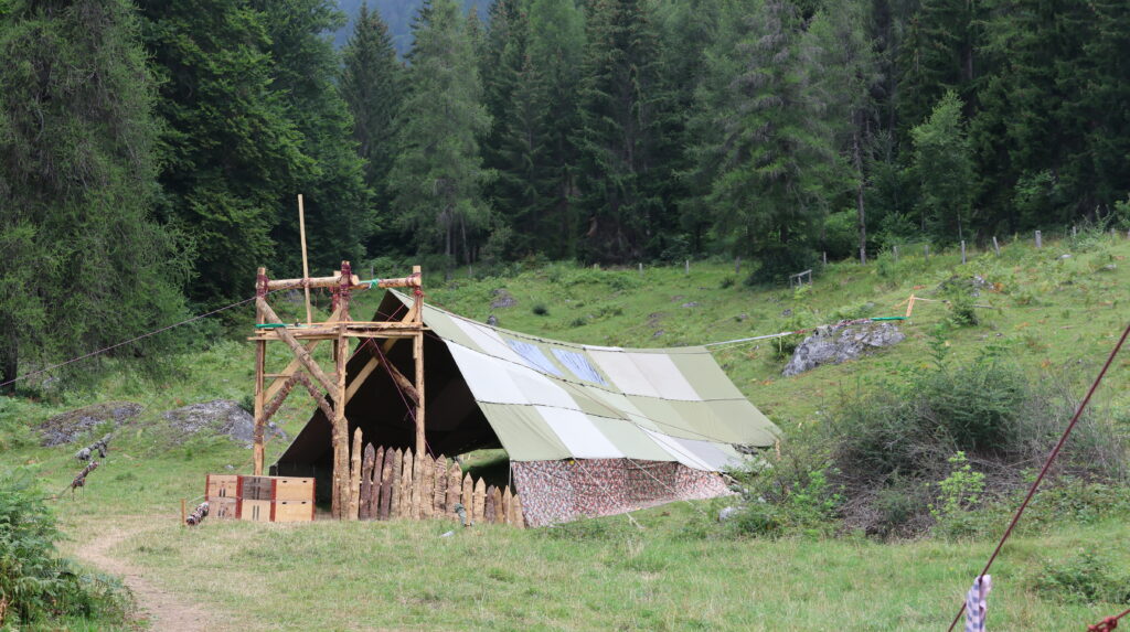 Lagerbauten und Blachenzelte auf einer Wiese am Waldrand.